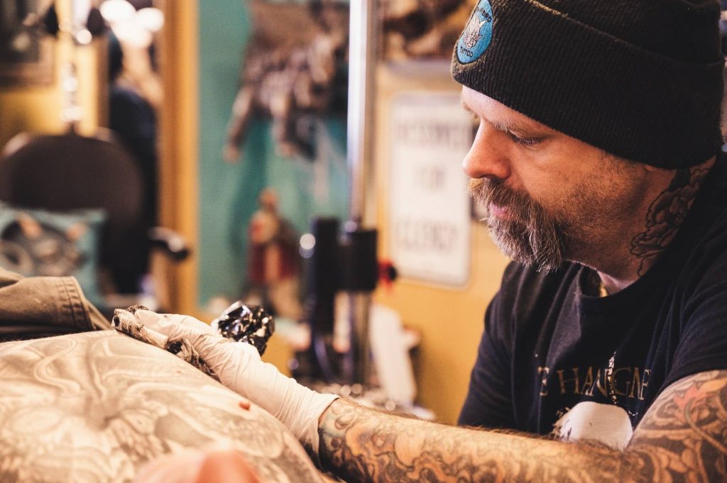 An image of a bearded and tattooed tattoo artist working on a tattoo, showcasing their expertise and skill in the art of tattooing, as they carefully create a design on a client's skin.