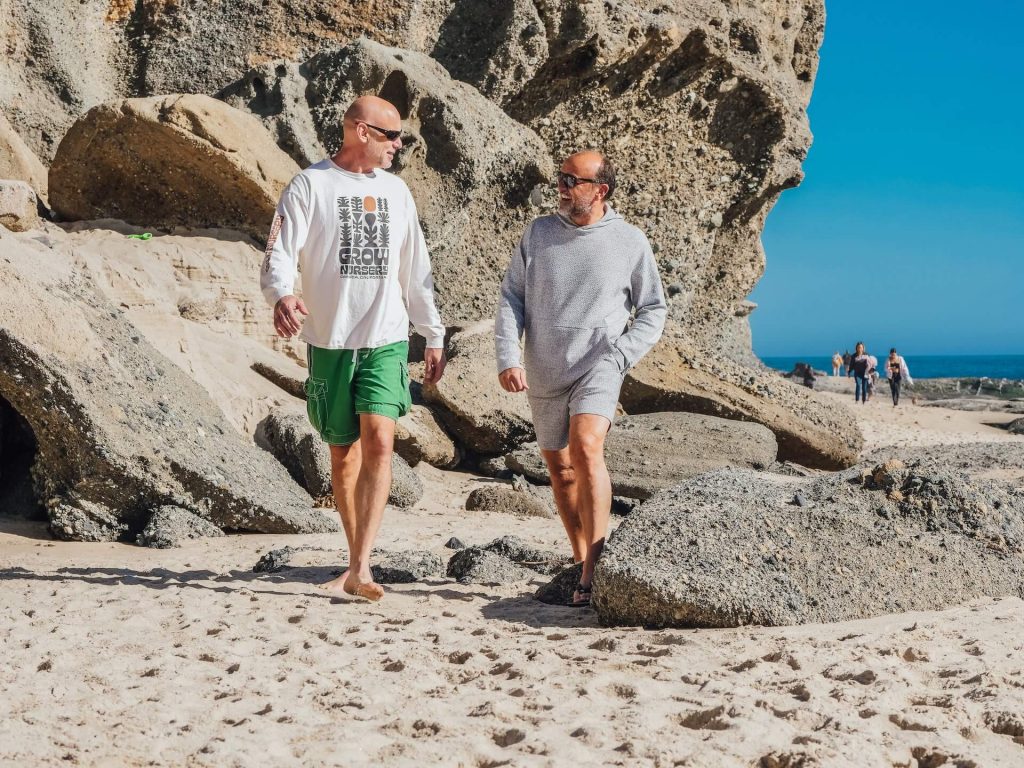 An image of two men strolling along the beach, both dressed in shorts and long-sleeve shirts, enjoying a leisurely walk by the seaside, showcasing a comfortable and relaxed beachwear style.